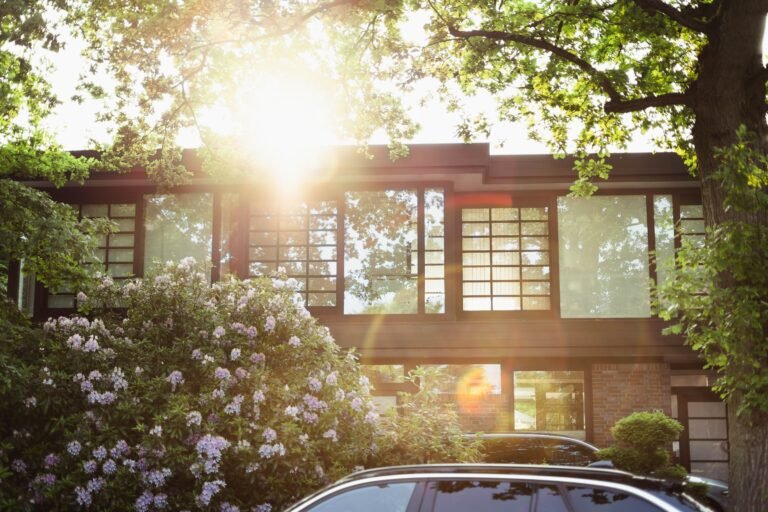 A sun-dappled modern house with large windows, garden, and car parked in front.